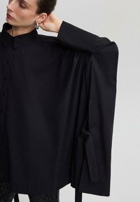 Czarna koszula z kołnierzykiem, zapinana na guziki z oversize'owymi rękawami i akcentami w postaci krawatów przy mankietach, wykonana z gładkiego, lekkiego materiału.