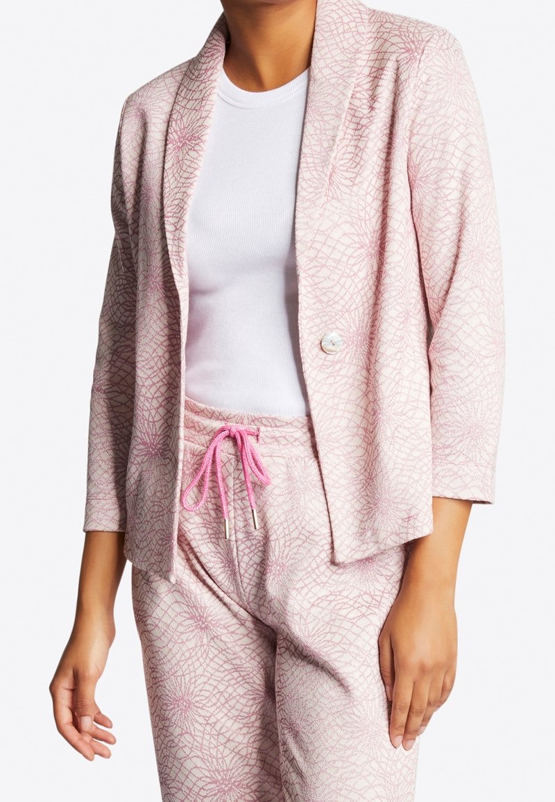 Femme portant un blazer rose clair à motif et un pantalon assorti avec un cordon rose, associé à un t-shirt blanc uni côtelé.