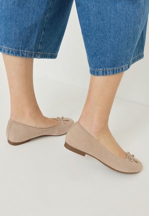 Pieds portant des ballerines beiges avec petits nœuds, assorties à un jean bleu court, debout sur un sol blanc.