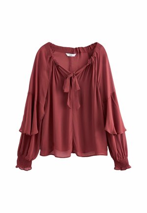 Femme portant un chemisier bordeaux ample et un jean bleu large avec des bottines marron à talons, debout devant un fond texturé uni.