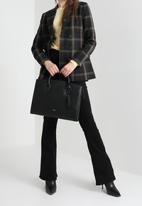 Sac à main structuré noir avec deux poignées et détail pompon, fabriqué en matériau lisse. Porté avec un blazer à carreaux et un pantalon noir évasé.