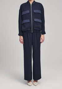 Marinblå zip-up bomberjacka med fyra matchande fickor, i kombination med löst sittande marinblå byxor och beigea ballerinas. Slät tygtextur.