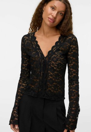 Une femme aux cheveux bouclés porte un haut en dentelle noire avec un décolleté festonné. Elle pose contre un fond blanc, dégageant une expression confiante.