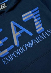 Felpa con cappuccio blu navy con grandi grafiche di lettere blu e beige "EA7" e "EMPORIO ARMANI" con logo. Texture morbida, stile casual.
