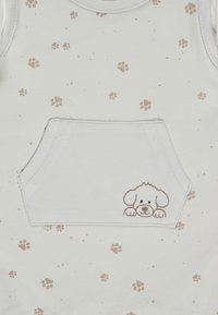 Weißes Babybekleidungsstück mit hellbraunen Pfotenabdrücken und einer vorderen Tasche, die ein kleines gesticktes Welpen-Gesicht zeigt, das über den Rand schaut.