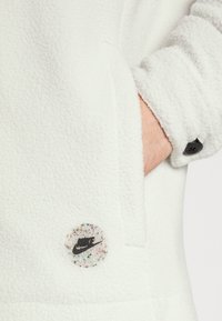 Handen i fickan på en vit fleecejacka med svarta knappärmar och en flerfärgad prickig Nike-logotyp nära fickan.