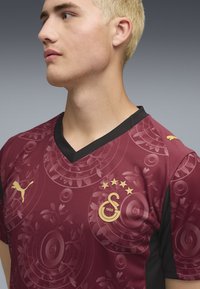 Kastanienbraunes Sporttrikot mit V-Ausschnitt, schwarzen Akzenten, goldenem Logo und Sternen sowie einem strukturierten Blumenmuster auf dem Stoff.