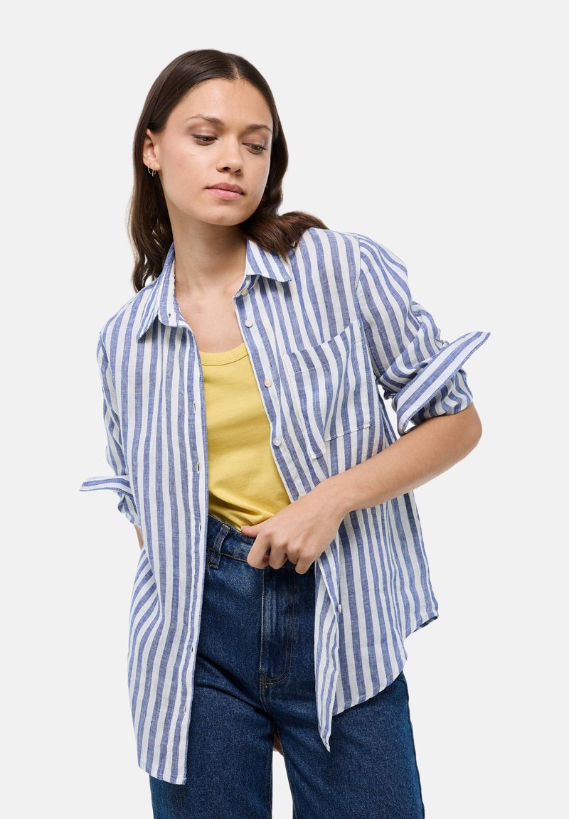 Chemise à rayures bleues et blanches, en tissu léger. Dotée de manches retroussables et d'une poche à rabat, portée par-dessus un haut jaune.