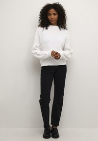 Maglione bianco a coste con maniche a sbuffo testurizzate, abbinato a pantaloni neri a gamba dritta e scarpe lucide nere. Design semplice e moderno.