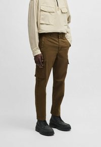 Bruine cargobroek van stevig materiaal, met zijzakken, een taps toelopend ontwerp en gecombineerd met zwarte veterschoenen.
