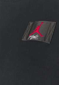 Czarna koszulka sportowa z teksturowanej tkaniny, z prostokątną naszywką przedstawiającą czerwone logo Jumpman oraz napis "Flight" w białym kursywie.