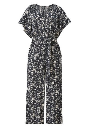 Žena oblečená v černo-bílém květovaném overal s páskem a bílými teniskami, stojící před jednobarevným pozadím.