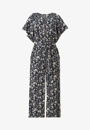 Mujer con un mono floral en blanco y negro con cinturón de lazo y zapatillas blancas, de pie frente a un fondo liso.