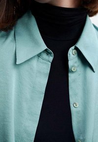Lys teal skjorte med knapper og krave, med en glat tekstur og matchende knapper, lagd over en sort turtleneck.