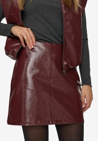 Donna che indossa una gonna in pelle bordeaux e un gilet sopra un maglione a maniche lunghe grigio scuro a coste, con collant neri visibili sotto la gonna.
