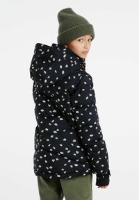 Zwarte pufferjack met een wit bloemenpatroon, voorzien van een capuchon en elastische manchetten, gecombineerd met een groene gebreide beanie en olijfgroene broek.