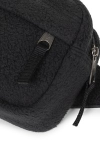 Bolsa de tela negra con textura y forma redondeada, con cierre de cremallera y una correa tejida. El material parece suave y peludo.