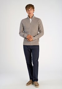 Beige zip-up tröja med ett strukturerat diamantmönster, i kombination med marinblå byxor och bruna slip-on skor, ställd mot en neutral bakgrund.