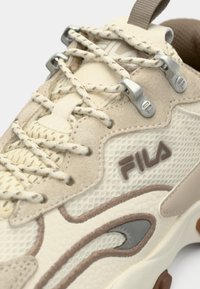 Μπεζ αθλητικό παπούτσι με υφή από δέρμα και δίχτυ στο πάνω μέρος, αντιθετικές καφέ λεπτομέρειες, μεταλλικά αυτιά κορδονιών και ξεχωριστή απεικόνιση λογότυπου.