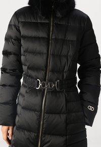 Abrigo de invierno negro acolchado con cuello de piel, cremallera frontal y cinturón con hebilla plateada con la letra "M" en la cintura.