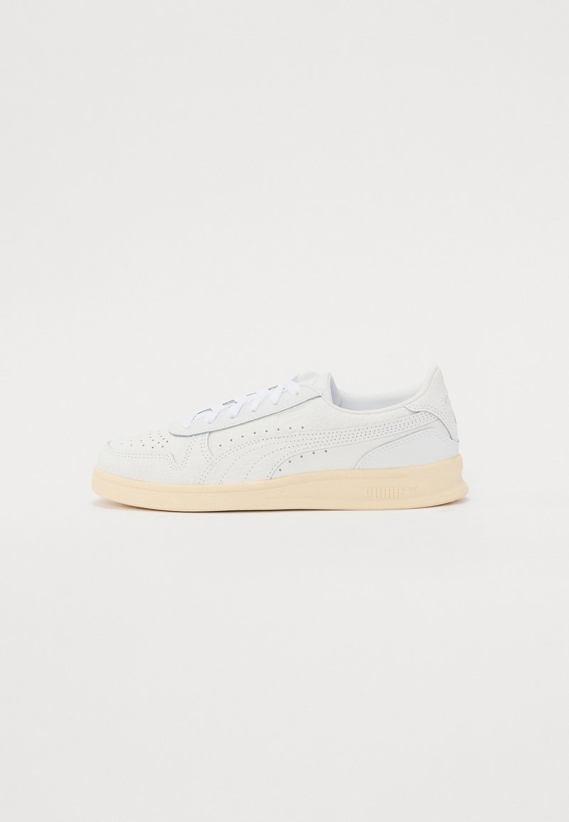 Baskets blanches basses avec des détails perforés, des lacets blancs et une semelle beige, présentées de profil sur un fond uni.