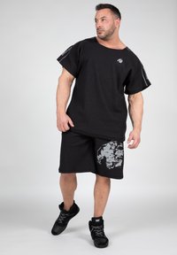 Zwarte ribbed shirt met korte mouwen en reflecterende details en logo, gecombineerd met zwarte shorts met een grijze grafische print, gedragen met zwarte sneakers.