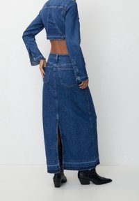 Chaqueta de mezclilla con mangas largas, de largo corto y dobladillo sin acabar, combinada con una falda larga de mezclilla con aberturas laterales y dos bolsillos traseros.