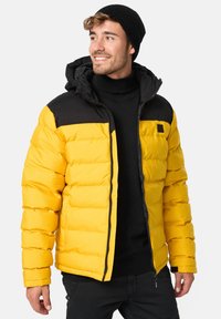 Gul pufferjakke med en sort, polstret skulderparti. Har hætte, lynlås fortil og ribbede manchetter. Glat tekstur og quiltet design.