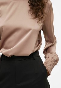 Žena na sebe má béžovú saténovú blúzku so smockovanými manžetami, zasunutú do čiernych elegantných nohavíc so zvýšeným pásom, pravá ruka v vrecku.