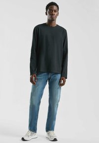 Zwarte lange mouwen shirt, blauwe klassieke jeans, en witte sneakers met gaasaccenten. Model staat tegen een lichte achtergrond.