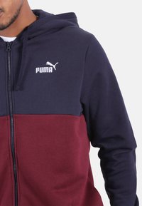 Felpa con cappuccio e zip Puma con sezione superiore blu navy e sezione inferiore bordeaux. Presenta un cappuccio, una cerniera frontale e un logo bianco sul petto. Materiale in misto cotone.