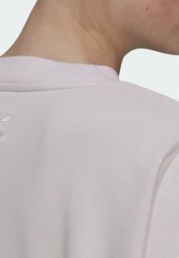 Bakifrån syns en person som bär en ljusrosa Adidas-tröja med ribbad krage och en liten broderad logotyp nära halslinningen.