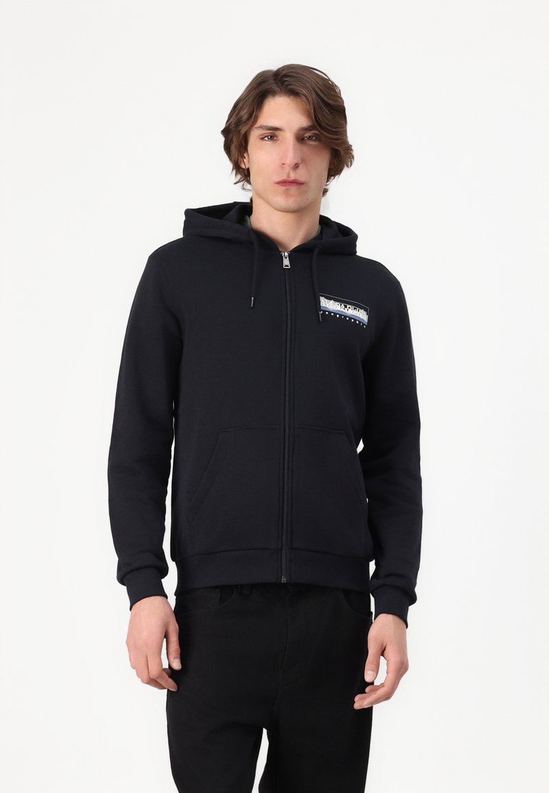 Młody mężczyzna w czarnej bluzie z zamkiem i kieszeniami z przodu, z logo, stoi na tle gładkiej białej ściany.