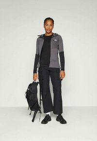 Pilko spalvos zip-up striukė su kapišonu, juoda marškinėliai, cargo kelnės ir juodi batai. Modelis laiko juodą kuprinę, stovi prieš neutralią fono.