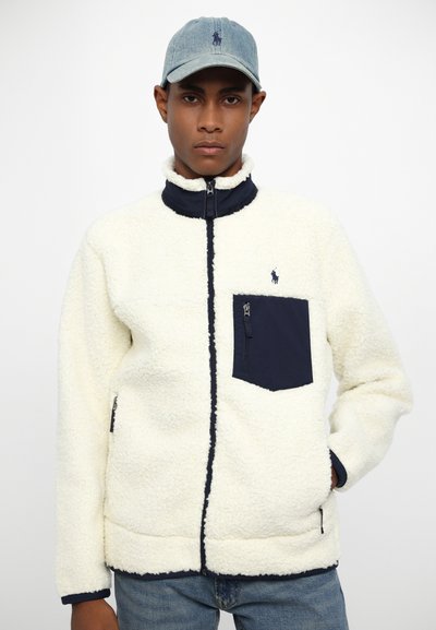 White fleece jacket with navy accents, zippered pocket, and standing collar. Worn with a light blue baseball cap.