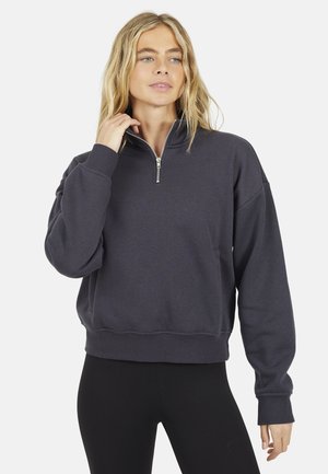 Donna con capelli biondi lunghi che indossa una felpa grigio scuro con mezza cerniera e leggings neri, in posa con una mano vicino al colletto.