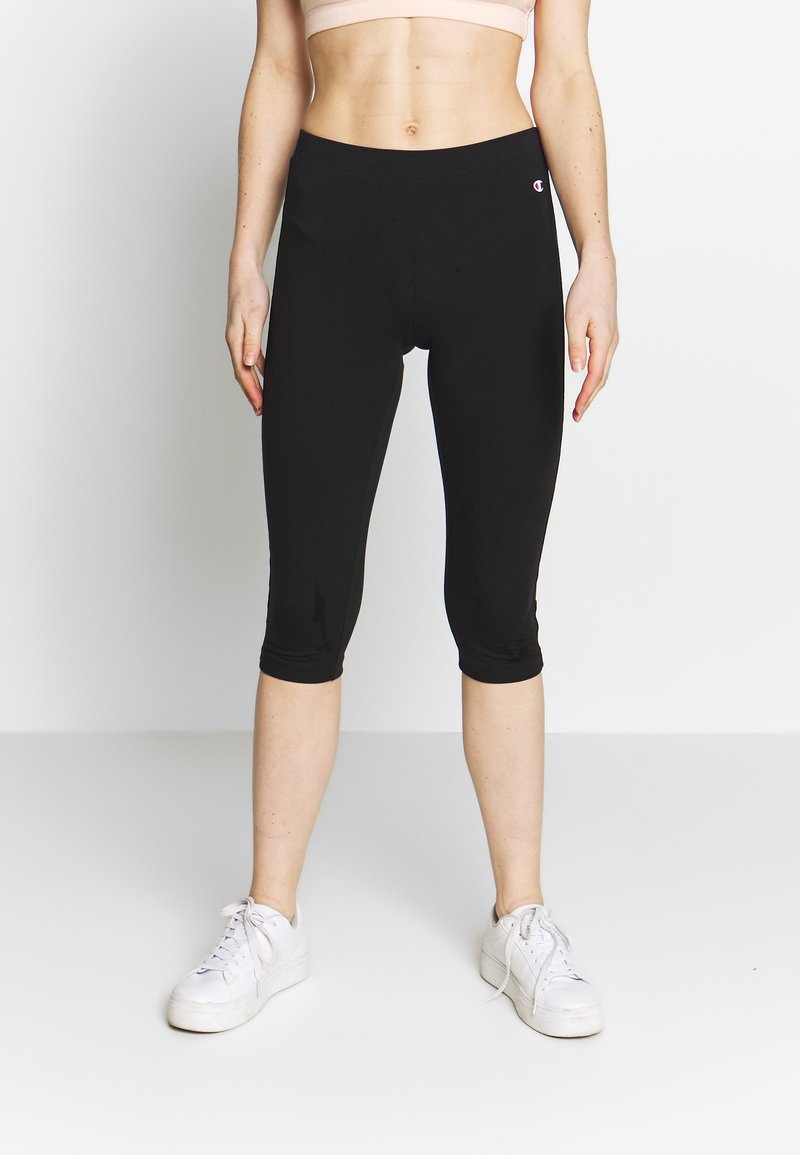 Kvinna som bär svarta korta leggings och vita sneakers stående mot en enkel vit bakgrund.