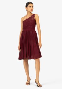 Bordeaux een-schouder jurk met kant bodice, geplooide rok en aangesloten taille. Gecombineerd met zwarte spiraal hak sandalen.