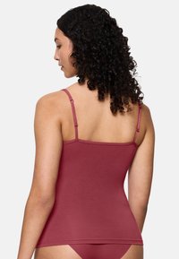 Kobieta z kręconymi ciemnymi włosami, ubrana w dopasowany bezrękawnik w kolorze bordowym oraz dopasowaną bieliznę, pokazana z tyłu na białym tle.