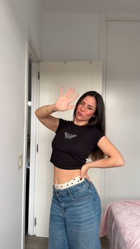 Svart avkortet t-skjorte med vinge-logo, høyvannede lyseblå denimjeans med prikkete midjebånd, stående i et lyst rom.