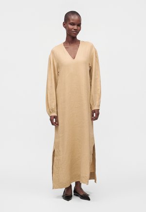 Frau trägt ein langes beiges Kleid mit langen Ärmeln, seitlichen Schlitzen und V-Ausschnitt, kombiniert mit schwarzen spitzen Ballerinas, steht vor weißem Hintergrund.