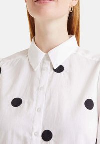 Camisa blanca de botones con cuello, adornada con grandes puntos bordados en negro. La tela parece ligera y texturizada.