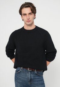 Pulover negru tricotat cu gât rotund, mâneci raglan și textură tip rețea, asortat cu blugi albaștri și o curea maro împletită.