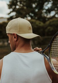 Beige sapka perforált oldalakkal és szilárd elülső résszel, logóval ellátott folttal. Világoskék, bordázott ujjatlan felsővel viselve, teniszkészletet tartva.
