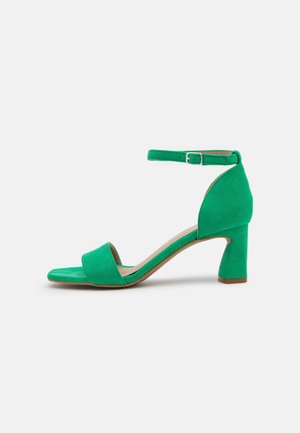 Sandalo verde in suede con punta aperta, cinturino alla caviglia e tacco a blocco su uno sfondo chiaro e semplice.