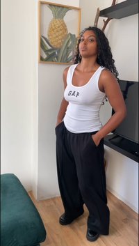 Top tank bianco con la scritta "GAP" in lettere nere, abbinato a pantaloni neri larghi. Il modello sta in piedi con le mani nelle tasche, su uno sfondo neutro.