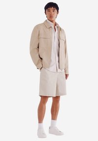 Beżowa lekka kurtka z dwoma kieszeniami na piersi, w połączeniu z białą koszulą i beżowymi szortami. Outfit uzupełniają białe tenisówki.