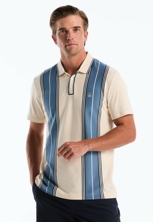 Homme portant un polo crème à rayures verticales bleues et un pantalon bleu marine, debout devant un fond blanc uni.