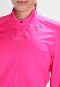 Veste zippée rose réalisée en tissu lisse et léger. Elle dispose d'un col haut, d'un logo adidas gris et d'une fermeture éclair à l'avant.