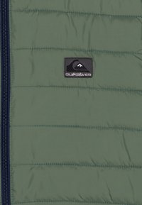 Quiksilver polstret vest i olivengrøn med vandret quiltede syninger, marineblå lynlås og et sort logo-patch på brystet.